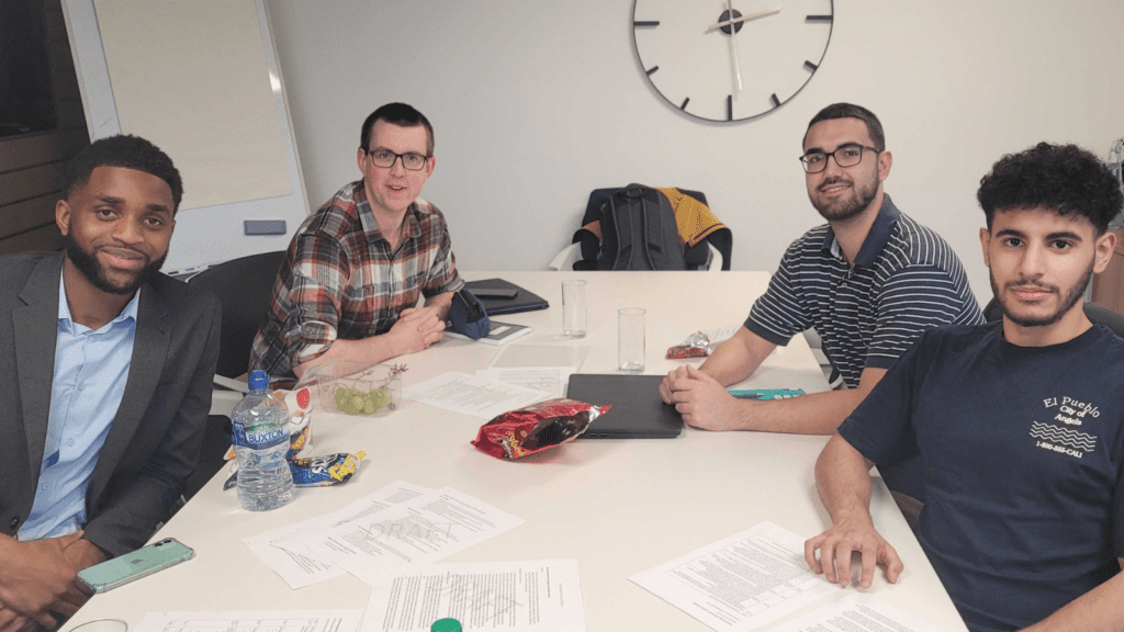 Four young men are gathered around a table in a meeting room, looking towards the camera