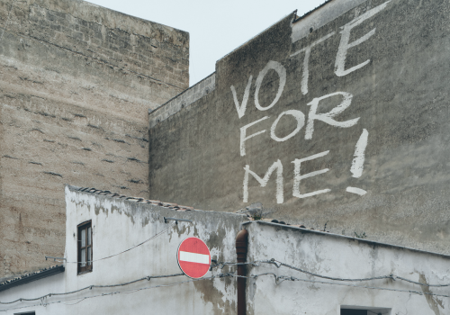 Graffiti on the outside of a run-down building reads "Vote for me!" There is a prominent road sign meaning no entry. The building could be part of the prison estate.